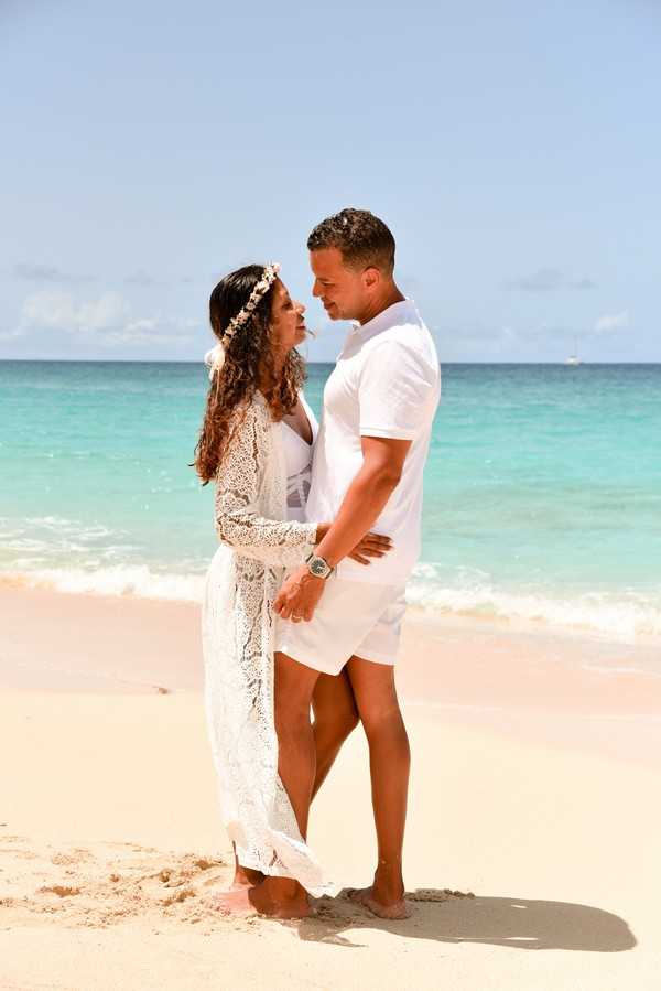 A couple poses together on a sandy beach in what appears to be a wedding portrait, standing barefoot at the water's edge and facing each other closely. The bride wears a white lace long-sleeve dress with a floral headband in her loose curly hair, while the groom wears an all-white short-sleeve shirt and shorts with a large-face watch. The setting is a tropical beach with turquoise water, styled in a relaxed boho aesthetic with no additional decor elements. The shot is a full-length portrait taken at mid-distance.