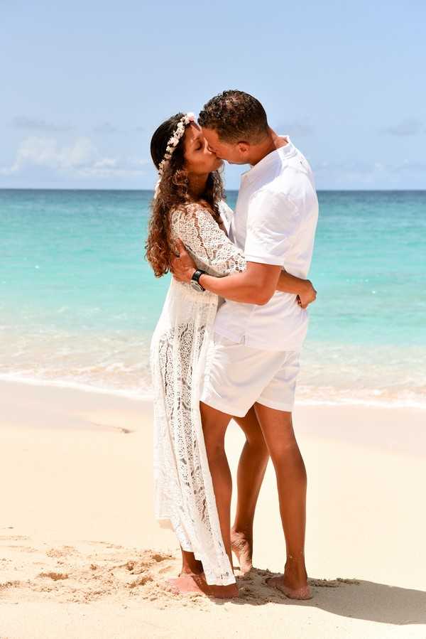A couple shares a kiss during a beach portrait session, standing barefoot on white sand at the water's edge with turquoise ocean water visible behind them. The bride wears a long white crochet lace dress with three-quarter sleeves and a floral crown with small pale pink and white blooms in her curly hair. The groom is dressed in an all-white outfit consisting of a short-sleeve button-up shirt and shorts, wearing a dark watch. The styling is relaxed boho beach with an all-white color palette for both outfits. The shot is a full-length portrait taken in natural daylight.
