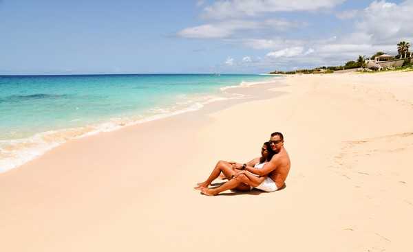 This image does not appear to be a wedding photograph. It shows a couple in casual beach attire — the woman in a dark bikini and the man shirtless in white shorts — sitting together on a wide, empty sandy beach with turquoise water in the background. There are no wedding-related elements such as a dress, suit, floral arrangements, or ceremony details visible. Wide-shot composition with the couple positioned in the lower-center of the frame.