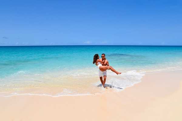 A couple poses on a tropical beach at the water's edge, with the groom lifting the bride in his arms as a gentle wave washes around their feet. The setting is an outdoor beach location with vivid turquoise water and white sand. The bride wears a short white dress, while the groom is shirtless in white shorts, suggesting an informal, relaxed beach wedding aesthetic. The shot is a wide portrait capturing both figures against the expanse of ocean and clear blue sky.