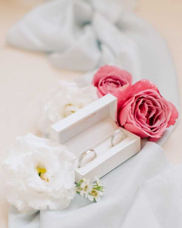 A close-up detail shot of two wedding bands displayed in an open white velvet ring box, placed on soft pale blue-grey draped fabric. The box contains one wider plain platinum band and one thinner diamond-set band. The arrangement is styled with coral-pink garden roses, white lisianthus blooms, and small white filler flowers surrounding the box. The overall color palette is soft and romantic, combining white, pale blue-grey, and coral-pink tones.