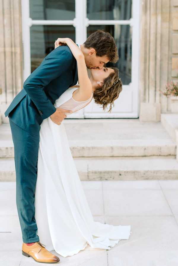 A couple portrait shot outdoors on the steps of a classical French building, featuring the groom dipping the bride as they kiss. The groom wears a fitted navy blue suit with tan oxford shoes and red socks, while the bride wears a minimalist white gown with a flowing skirt and a blush-toned bodice, accessorized with a delicate hair piece. The bride has short, wavy dark hair and the composition is a full-length portrait with the architectural facade and white French doors visible in the background. The styling is modern and understated with a classic French setting.