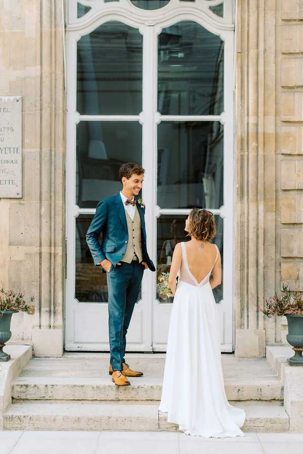 A couple portrait taken outdoors on the stone steps of a classical French building, identifiable by a plaque referencing Lafayette and a date of 1779, likely a historic hôtel particulier or town hall. The groom faces the camera smiling, wearing a teal blue suit with a beige waistcoat, mustard yellow Oxford shoes, and a floral bow tie with a small boutonnière. The bride stands with her back to the camera, wearing a white floor-length gown with a deep open back featuring delicate lace trim along the neckline, and she holds a small bouquet. The shot is a medium full-length portrait capturing both figures, with large white-painted French doors and ornate stone architecture as the backdrop. Potential venue feature image.