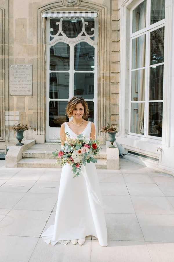 A bride stands alone in front of a classical French stone building, posing for a full-length portrait outdoors on a pale stone terrace. She wears a sleek, fitted white gown with a V-neckline and minimal embellishment, paired with white low-heeled shoes and a delicate pearl necklace, with a small hair accessory worn across her short, wavy dark hair. She holds a loosely arranged bouquet featuring ivory garden roses, deep red roses, blush pink ranunculus, and abundant eucalyptus greenery. The building behind her features ornate white-painted ironwork above a tall door, decorative stone carvings, and two dark urn-style planters flanking the entrance steps, giving the setting a classic Parisian aesthetic.