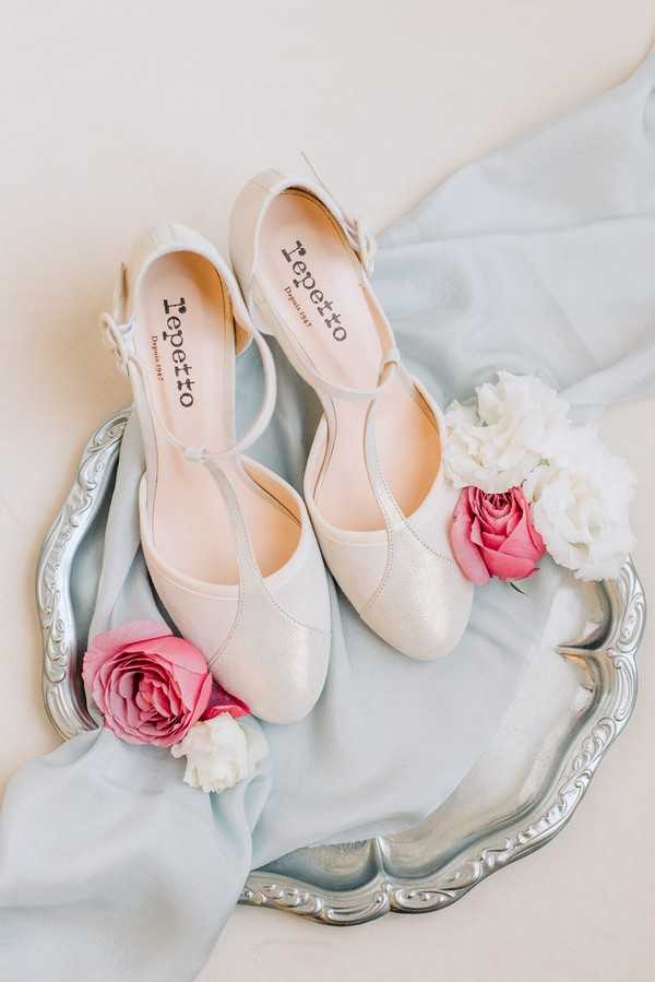 A flat-lay detail shot of a pair of Repetto brand ivory T-strap low-heeled bridal shoes displayed on a silver ornate tray. The shoes are styled with a pale blue silk ribbon draped underneath and surrounded by hot pink ranunculus blooms and white peony petals. The overall palette is soft and romantic, combining ivory, silver, pale blue, and bright pink accents.