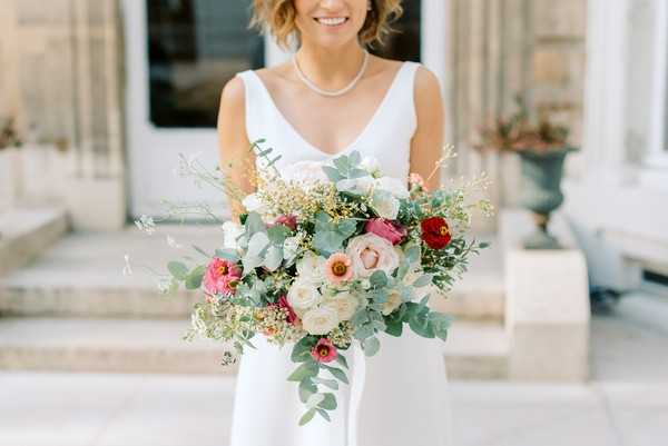 A bridal portrait close-up showing a bride from the chest down to mid-thigh, holding a large, loosely arranged bouquet in front of a classical stone building with columns and decorative urns visible in the background. The bride wears a sleeveless white gown with a V-neckline and a pearl necklace, with short wavy hair and a broad smile. The bouquet features blush and cream garden roses, deep red ranunculus, hot pink zinnias or ranunculus, ivory blooms, eucalyptus sprigs, and delicate golden filler flowers in an organic, unstructured style. The overall palette of the bouquet mixes warm reds and pinks with soft neutrals and silvery-green foliage.
