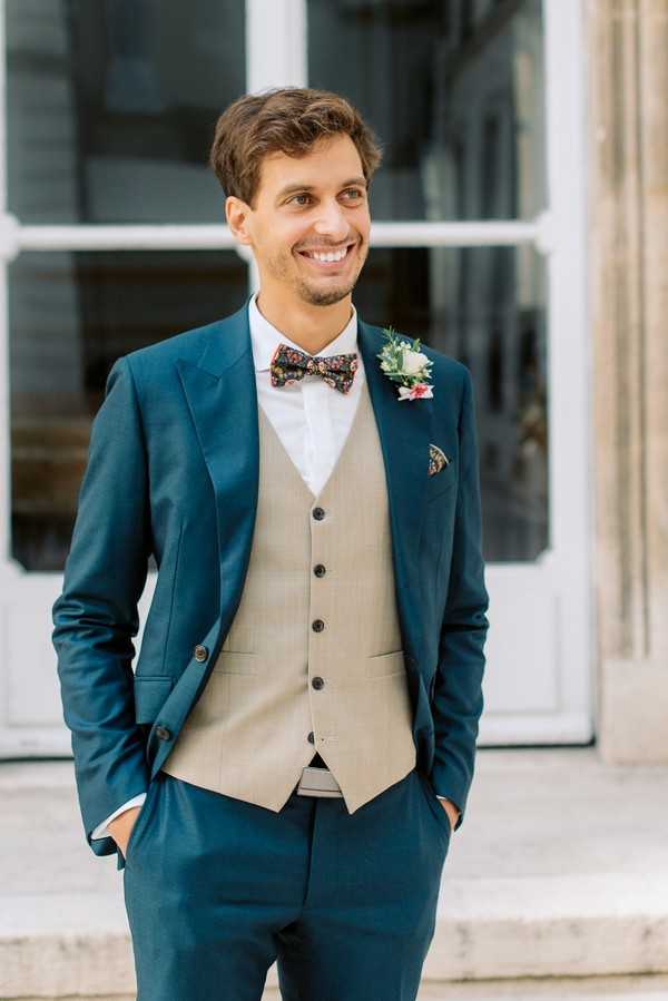 A portrait of a groom standing outdoors in front of a building with large white-framed windows. He is wearing a teal blue suit jacket and trousers paired with a contrasting tan/khaki waistcoat, a white dress shirt, and a dark floral-print bow tie with a matching floral pocket square. His boutonniere features a small white flower with red accents and greenery. He is smiling and looking slightly off-camera with his hands in his pockets. The overall styling is modern with eclectic, pattern-mixed accessories.
