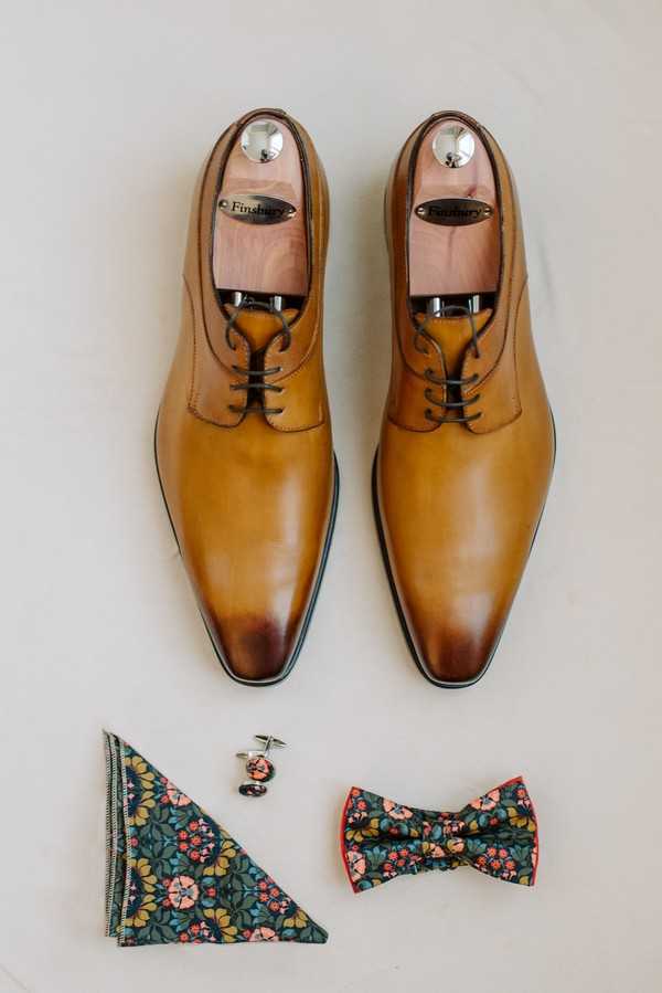 A flat lay detail shot of groom's accessories arranged on a light grey/white surface. The focal point is a pair of tan leather Finsbury oxford dress shoes with cedar shoe trees inserted and dark brown laces. Below the shoes are three coordinating accessories: a folded pocket square, a pair of cufflinks, and a bow tie — all in a matching dark teal Liberty-style floral print featuring coral, pink, and mustard yellow botanical motifs with a coral red border on the bow tie.
