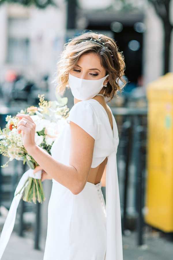 A portrait of a bride standing outdoors on an urban street, looking down at her bouquet. She wears a fitted white gown with a open back and cap sleeves, a matching white face mask, pearl earrings, and a delicate green leaf headpiece in her short wavy hair. She holds a bouquet of white garden roses, small orange accent blooms, eucalyptus, and fine white filler flowers tied with a white ribbon. The background is a blurred city streetscape with café chairs and a yellow post box visible, suggesting a French urban setting. The shot is a close-up portrait with a shallow depth of field and soft natural light.