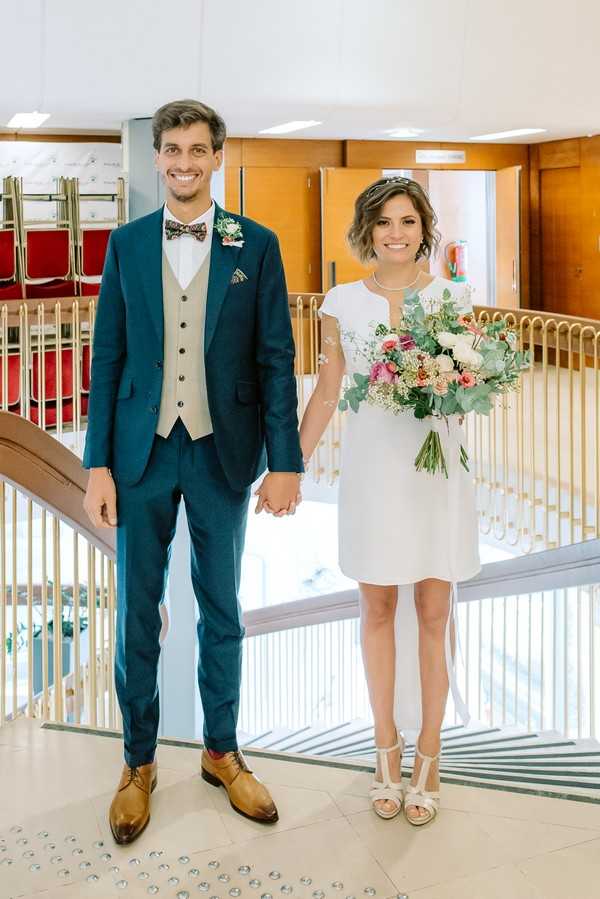 A couple poses hand-in-hand indoors in what appears to be a French civil ceremony venue (mairie), standing near a gold-railed staircase with red chairs visible in the background. The groom wears a teal two-piece suit with a beige waistcoat, a floral bow tie, a boutonnière of small white and pink blooms, and tan leather oxford shoes. The bride wears a short, cap-sleeve white shift dress with subtle embroidered detail, white t-strap heeled sandals, and carries a loose bouquet of ivory roses, coral and pink blooms, and eucalyptus foliage. The image is a full-length portrait shot with natural indoor lighting.