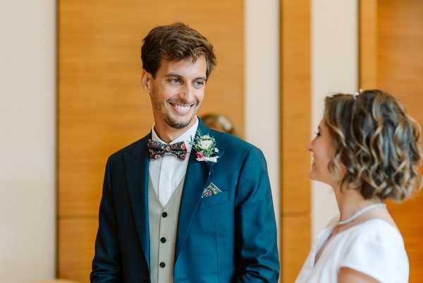 A close-up portrait of the groom and bride facing each other, both smiling, likely during the ceremony or a first-look moment indoors. The groom wears a teal blue suit jacket with a light grey waistcoat, a floral-print bow tie, a matching floral pocket square, and a boutonnière with small white and pink blooms. The bride, shown partially from behind, wears a white dress and has short wavy hair. The setting appears to be an indoor venue with warm wood-paneled walls in the background.