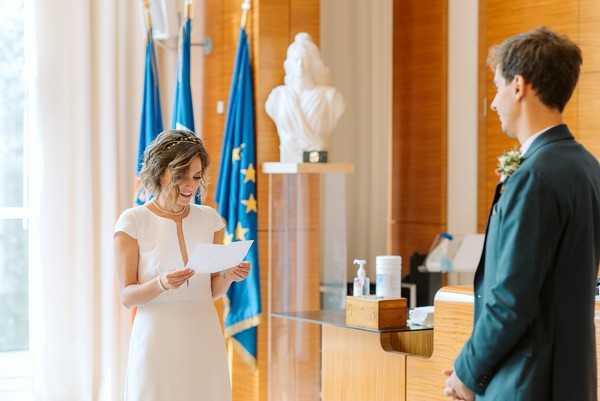 A civil wedding ceremony taking place inside a French town hall (mairie), with the bride reading from a piece of paper, likely her vows, while the groom stands facing her. The bride wears a simple short-sleeved white dress with a center neckline detail and a delicate headband, while the groom is dressed in a dark teal suit with a small floral boutonniere. The room features wood-paneled walls, a white marble bust on a pedestal, and French and European Union flags in the background, clearly identifying this as an official civil ceremony space. The image is a medium portrait-style shot with soft natural light coming from the left.