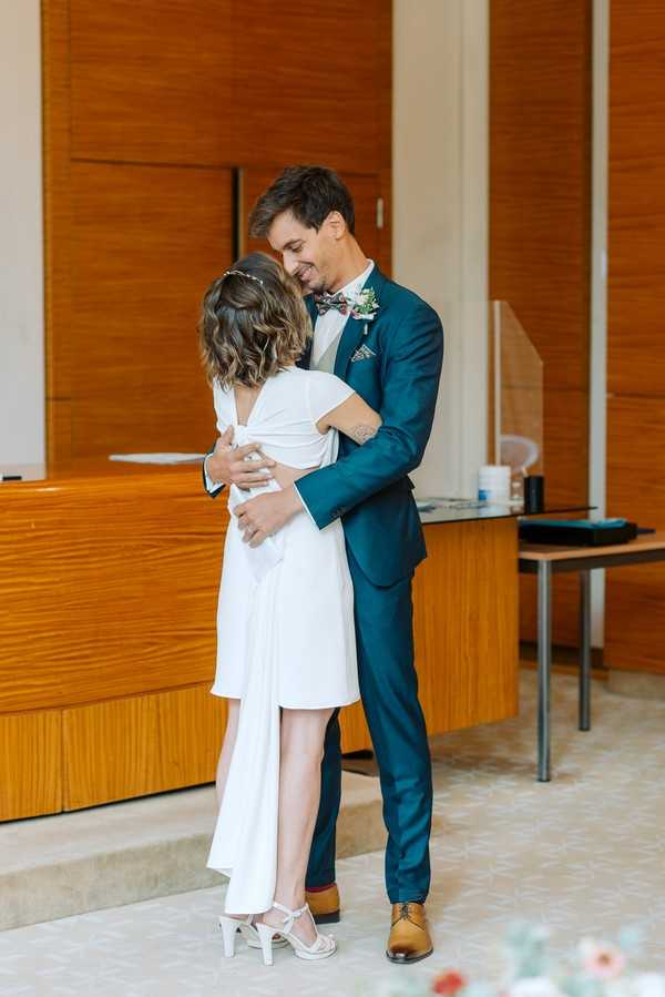 A couple embraces inside what appears to be a French civil registry office (mairie), likely just after completing their ceremony. The bride wears a short white dress with a low V-back, a small hair accessory, and white strappy heeled sandals, while the groom wears a teal/petrol blue suit with tan oxford shoes, a floral bow tie, and a small boutonniere with pink flowers. The setting is a modern interior with warm wood-paneled walls and a reception desk visible in the background. The shot is a medium full-length portrait capturing the couple from the front and back respectively, with the groom smiling as they hold each other.