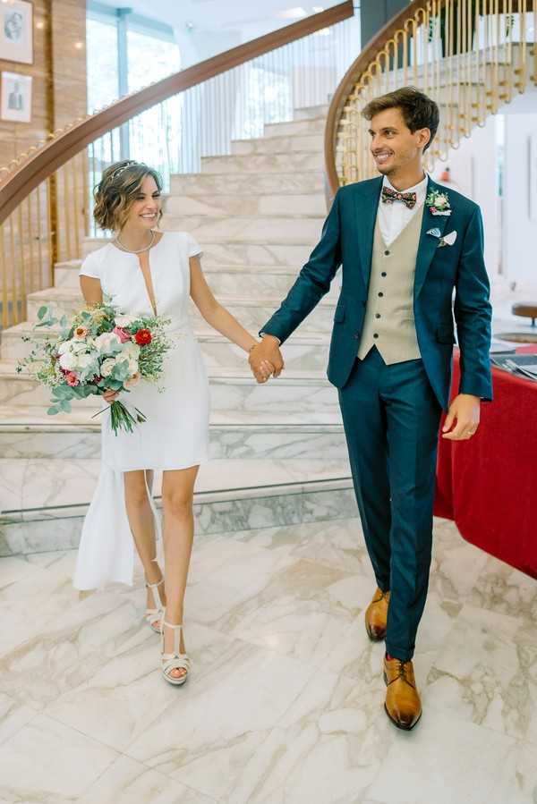 A bride and groom walk hand-in-hand down a grand curved marble staircase inside what appears to be a formal civic or hotel building, smiling at each other. The bride wears a short, cap-sleeve white dress with a deep keyhole neckline, white strappy heeled sandals, and a delicate headband, carrying a loose bouquet of white and red blooms with eucalyptus greenery. The groom wears a fitted navy blue suit with a beige waistcoat, floral bow tie, a small floral boutonniere, and tan leather oxford shoes. The setting features white marble floors and stairs with warm gold stair railings, giving the scene a modern classic feel. This is a full-length couple portrait shot taken indoors with bright natural light.