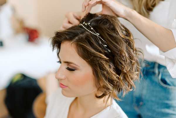 A close-up portrait shot of a bride during the getting-ready phase, with a stylist's hands placing a delicate crystal and leaf hair vine accessory into her short, wavy dark brunette hair with highlights. The bride is wearing a white robe and has finished makeup including defined brows, smoky eye shadow, and a coral-pink lip. The hair accessory features small pearl or crystal embellishments along a thin wire with leaf motifs. The background is softly blurred in a bright, light-filled indoor setting.