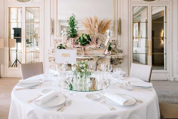 A detail shot of a round reception dining table set inside a classic French chateau ballroom with white paneled walls, ornate stone fireplace, and mirrored doors. The table is dressed in a white floor-length linen with white charger plates, white folded napkins, gold flatware, and multiple wine glasses per setting. The centerpiece features small glass bud vases with delicate white and green florals arranged on a round mirror base, alongside a gold-lettered table name card reading 'Paris.' The fireplace mantel in the background is decorated with pampas grass, small potted topiaries, and rose gold metallic vases, reinforcing a neutral, modern-classic decor palette of white, gold, and natural greens.