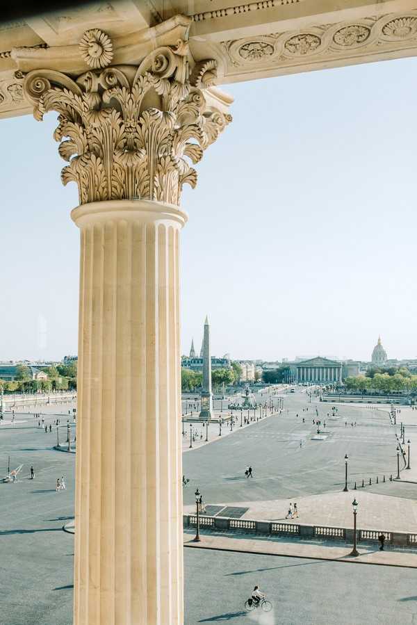 This image is not a wedding photograph but rather an architectural detail and cityscape shot taken from an elevated vantage point in Paris. The foreground features a large cream-colored Corinthian column with ornate acanthus leaf capital detail, framing a wide view of the Place de la Concorde below, with the Luxor Obelisk, the Madeleine church, and the Hôtel des Invalides dome visible in the background. A handful of pedestrians and a cyclist are visible in the plaza. The composition is a wide architectural shot using the column as a foreground framing element. Potential venue feature image.