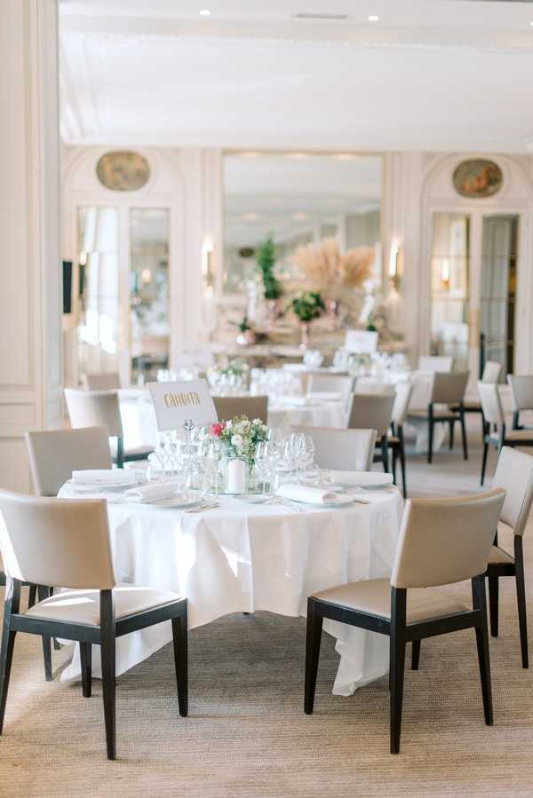 Indoor wedding reception setup inside a classic French ballroom or salon with white-paneled walls, ornate oval medallion reliefs, and mirrored panels with warm wall sconce lighting. The foreground features a round reception table dressed in a white floor-length linen, set with crystal wine glasses, white folded napkins, and a low centerpiece of white and blush florals with greenery, accompanied by a handwritten gold-lettered table name card. The chairs are modern dark-wood frames with taupe/greige upholstered seats and backs, mixing a classic venue with contemporary furniture. Multiple additional round tables are visible in the background, one featuring a tall arrangement of pampas grass and greenery in a blush-toned vase. The overall decor palette is white, ivory, and warm neutral tones with soft ambient lighting. Wide interior shot with no people present. Potential venue feature image.