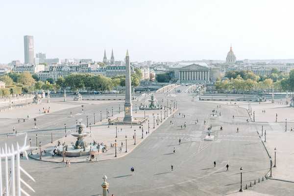 This wide aerial shot shows the Place de la Concorde in Paris, with the Luxor Obelisk and ornamental fountains visible in the foreground plaza, and landmarks including Les Invalides and the Assemblée Nationale visible in the background skyline. The image appears to be taken from an elevated position, possibly a rooftop or upper floor of a nearby building, with a decorative gold finial and white ironwork visible at the lower left edge of the frame. No wedding subjects, couple, or wedding-related decor are visible in this image — it functions purely as a wide establishing shot of a Paris cityscape. Potential venue feature image.