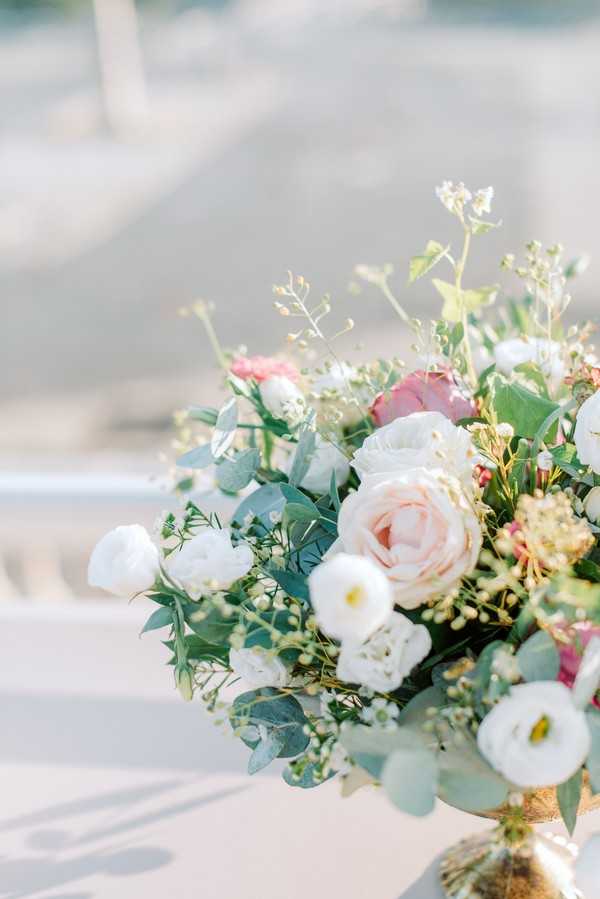 Close-up detail shot of a floral arrangement placed on what appears to be a white-draped surface at an outdoor wedding. The arrangement features blush pink garden roses, white lisianthus, dusty pink protea, small yellow wax flower clusters, eucalyptus foliage, and delicate wispy greenery, all set in a gold urn or vessel. The color palette is soft blush, white, and sage green, consistent with a romantic garden-style aesthetic. The background is heavily blurred, suggesting an open outdoor setting with bright natural light.