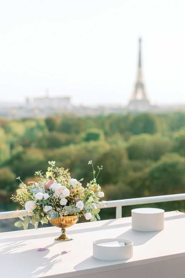 A detail close-up shot of a wedding table arrangement on an outdoor terrace in Paris, with the Eiffel Tower visible and softly blurred in the background. The foreground features a gold urn-style vase holding a low floral arrangement of blush pink garden roses, white lisianthus, and eucalyptus with delicate filler greenery, alongside two white cylindrical boxes likely containing rings or wedding stationery. The table surface is white, and a few scattered petals are visible. The overall decor palette is soft and romantic with gold, blush, and white tones in a classic Parisian style. Potential venue feature image.