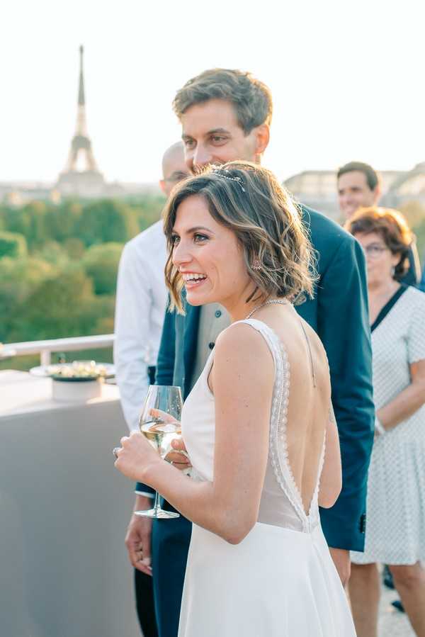 A cocktail hour scene on an outdoor rooftop terrace in Paris, with the Eiffel Tower visible in the background during golden hour. The bride stands in the foreground holding a glass of white wine or champagne, wearing a white low-back dress with lace trim detailing along the open back, short wavy hair adorned with a delicate hair accessory, and a diamond necklace. The groom stands directly behind her in a navy blue suit, and several guests are visible in the background, including a woman in a white polka-dot dress. The image is a medium portrait shot with warm evening light.