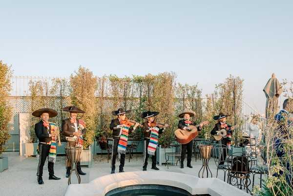 A mariachi band of seven musicians performs outdoors on a rooftop terrace during what appears to be a cocktail hour or reception entertainment set. The musicians are dressed in traditional charro suits — black embroidered jackets and trousers — with large black sombreros and colorful serape-style sashes in red, green, orange, and blue stripes. The band includes violinists, a guitarist, and a vocalist, performing in a line formation. The terrace setting features low white curved seating around a small water feature, ornate black wrought-iron café chairs and tables, and tall green hedged trellises as a backdrop. A wide-shot composition captures the full group against a pale evening sky, with a partially visible guest in a blue outfit on the right edge of the frame.