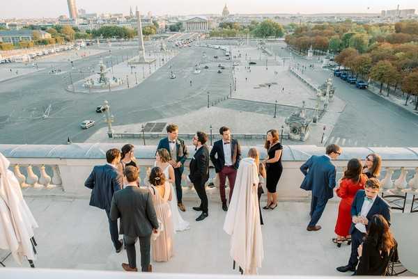 A cocktail hour or reception gathering takes place on a rooftop terrace with a white stone balustrade, overlooking the Place de la Concorde in Paris, with the Panthéon dome visible in the background. Approximately 15 guests and the wedding couple are mingling on the terrace, with the bride wearing a white gown and the groom in a dark suit with a bow tie. Guests are dressed in cocktail attire including a blush/champagne bridesmaid dress, a red dress, and various dark suits. The image is a wide aerial shot taken from above, capturing both the social gathering in the foreground and the sweeping Parisian cityscape beyond. Potential venue feature image.