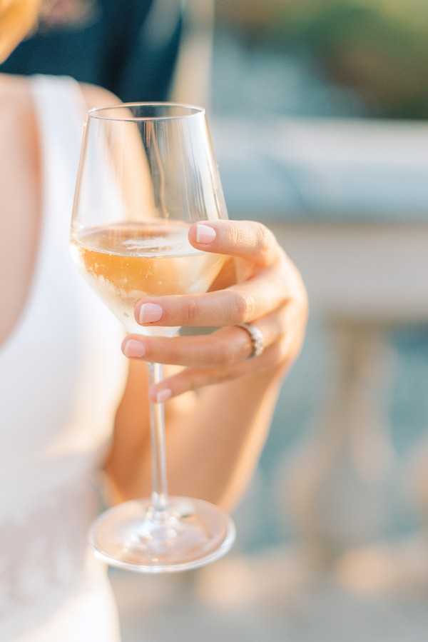 Close-up detail shot of a woman's hand holding a wine glass filled with champagne or white wine, likely during a cocktail hour or reception. The subject wears a white dress visible at the edge of the frame and a delicate ring on her finger, with pale pink manicured nails. The background is softly blurred but suggests an outdoor terrace setting with what appears to be a stone balustrade, bathed in warm golden light.
