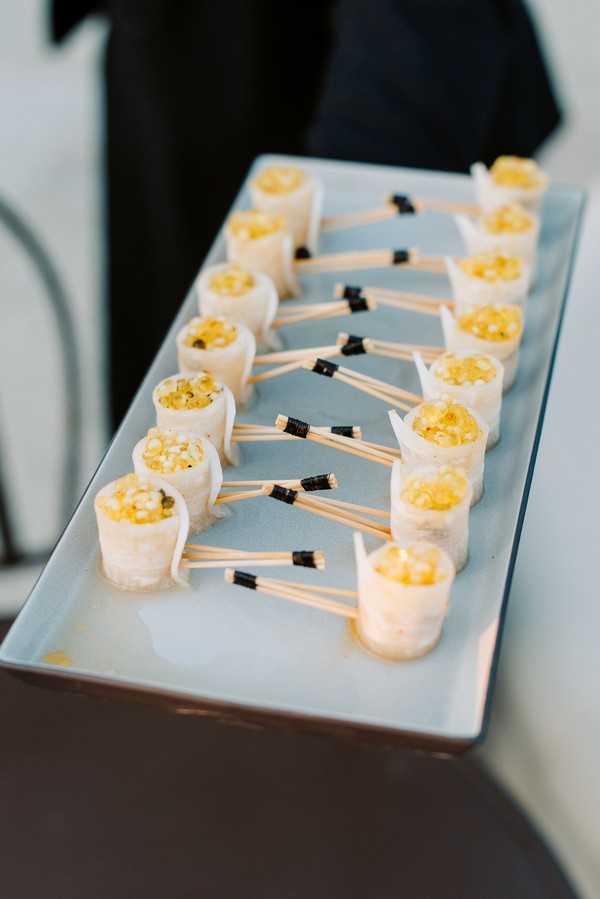 Close-up detail shot of cocktail hour canapés arranged on a rectangular pale blue-grey serving tray, held by a person dressed in black visible in the blurred background. Approximately fourteen small rice paper rolls filled with a golden-yellow topping — likely a corn or citrus-based garnish — are lined up in two rows and each skewered with a thin wooden cocktail pick banded with a small black tie detail. The presentation style is modern and structured, suggesting a contemporary catering aesthetic.