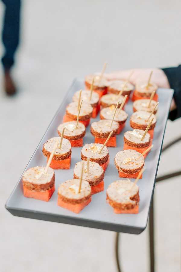 Close-up detail shot of a cocktail hour canapé service, showing a server holding a rectangular white ceramic tray lined with approximately 18 bite-sized appetizers. Each canapé consists of a cube of fresh watermelon topped with a round slice of spiced, herb-crusted cheese log garnished with gold leaf flakes and skewered with a bamboo cocktail pick. The tray is held outdoors against a neutral concrete background, with a guest's legs in navy trousers and brown shoes visible in the blurred background. The presentation reflects a modern, upscale catering style with a pop of warm orange from the watermelon against the pale white tray.