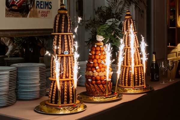 A reception detail shot showing three croquembouche wedding cakes displayed on a buffet table, each fitted with lit sparklers that are actively sparkling. The two outer croquembouches are tall and slender cone-shaped towers built with cream puffs and caramel, while the central one is slightly shorter and wider with a more rounded silhouette and visible golden-caramel profiteroles. All three are presented on gold circular bases. To the left, stacks of grey-rimmed plates are visible, and to the right, a champagne bottle and glasses can be seen. A vintage Reims motor racing poster dated 5 Juillet 1959 is mounted on the wall in the background, suggesting an indoor restaurant or bistro-style reception venue with warm, dim ambient lighting.