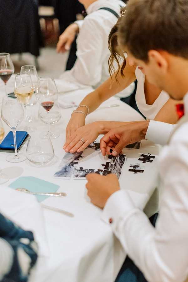 A close-up detail shot taken during a wedding reception, showing two guests seated at a white-linen-covered table assembling a black-and-white photo puzzle as a table activity. The woman wears a white dress and a pearl bracelet with a ring visible on her hand, while the man beside her wears a white dress shirt with cufflinks. The table is set with multiple wine glasses containing white and red wine, a blue phone or card holder, light blue place cards, and a pen. The puzzle pieces are spread across what appears to be a black-and-white photograph of the couple. The composition is a close overhead angle focusing on the hands and table details.