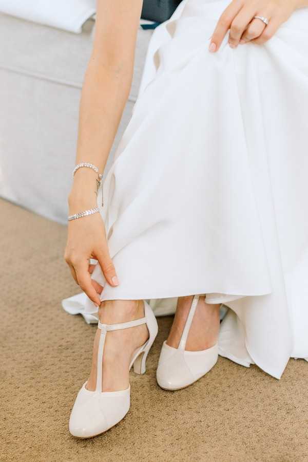 A close-up detail shot of a bride putting on her shoes during the getting-ready phase of the wedding day. She is wearing ivory patent leather T-strap heeled pumps and a white voluminous gown with a smooth satin-like fabric. Her accessories include a delicate diamond tennis bracelet on her wrist and an engagement ring on her finger. The image is cropped at mid-torso, focusing on her hands, shoes, and the lower portion of her dress.
