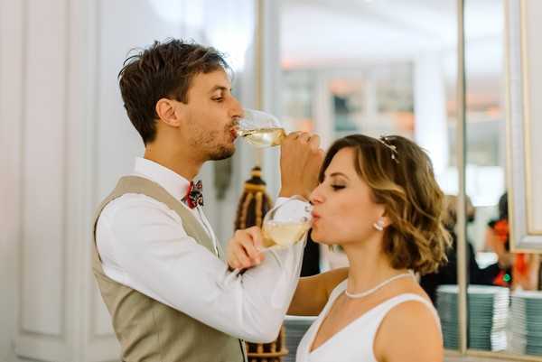 The bride and groom are performing a champagne coupe toast, drinking from interlinked glasses in the traditional crossed-arms style. The groom wears a tan/khaki vest over a white shirt with a burgundy floral bow tie, while the bride wears a white sleeveless dress with a pearl necklace and a delicate hair accessory. The setting appears to be an indoor reception space with large glass doors or windows visible in the background. This is a close-up portrait shot capturing the couple from the shoulders up mid-sip.