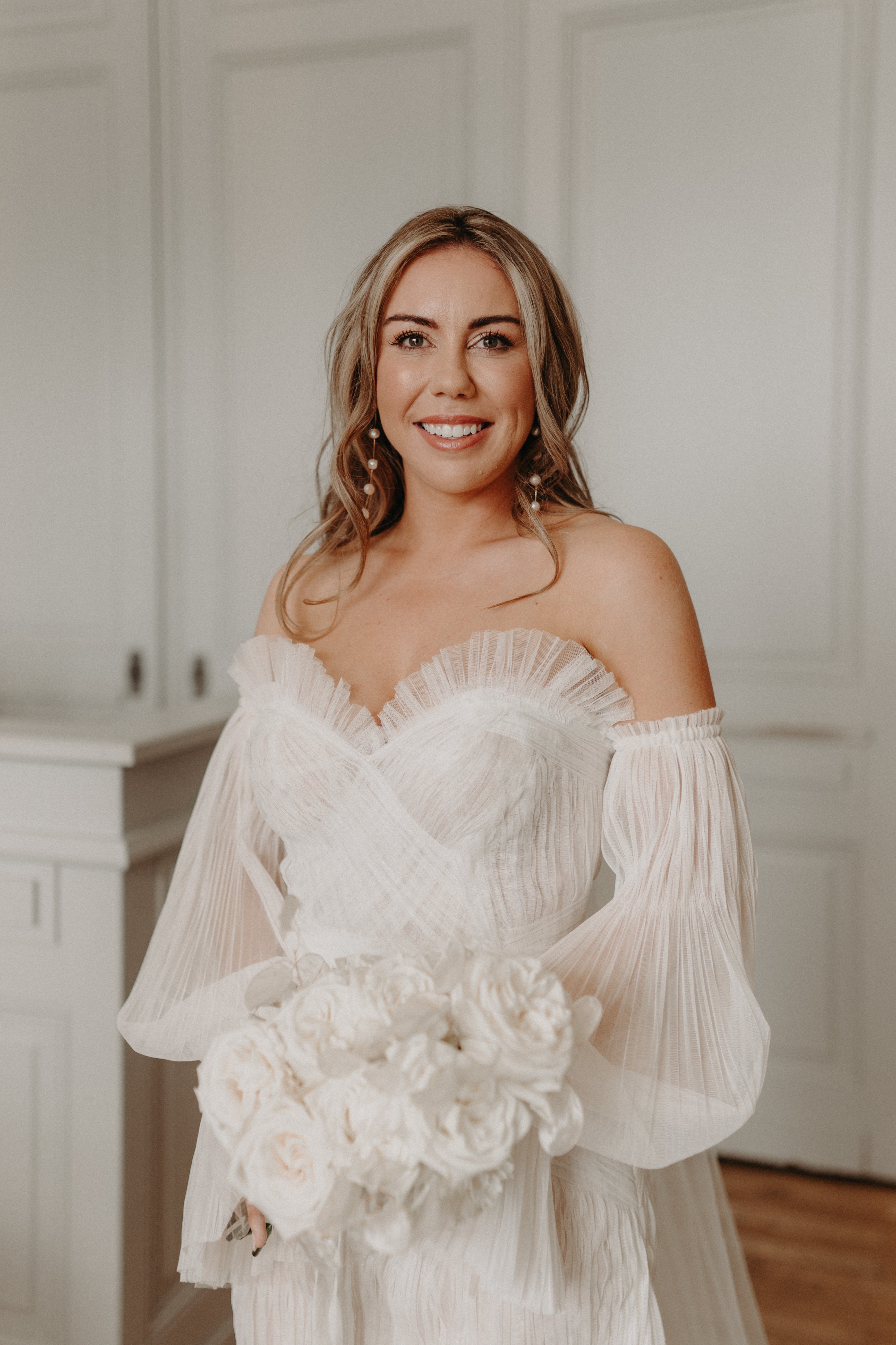 A bridal portrait taken indoors, showing the bride from approximately the waist up against a white panelled wall interior. The bride wears an off-the-shoulder white pleated tulle gown with detached billowing long sleeves and a sweetheart neckline with ruffled trim. She holds a compact, tightly arranged bouquet of white peonies and roses. Her accessories include dangling multi-pearl drop earrings, and her blonde hair is styled in loose waves. The composition is a close-up portrait shot with soft, natural indoor lighting.