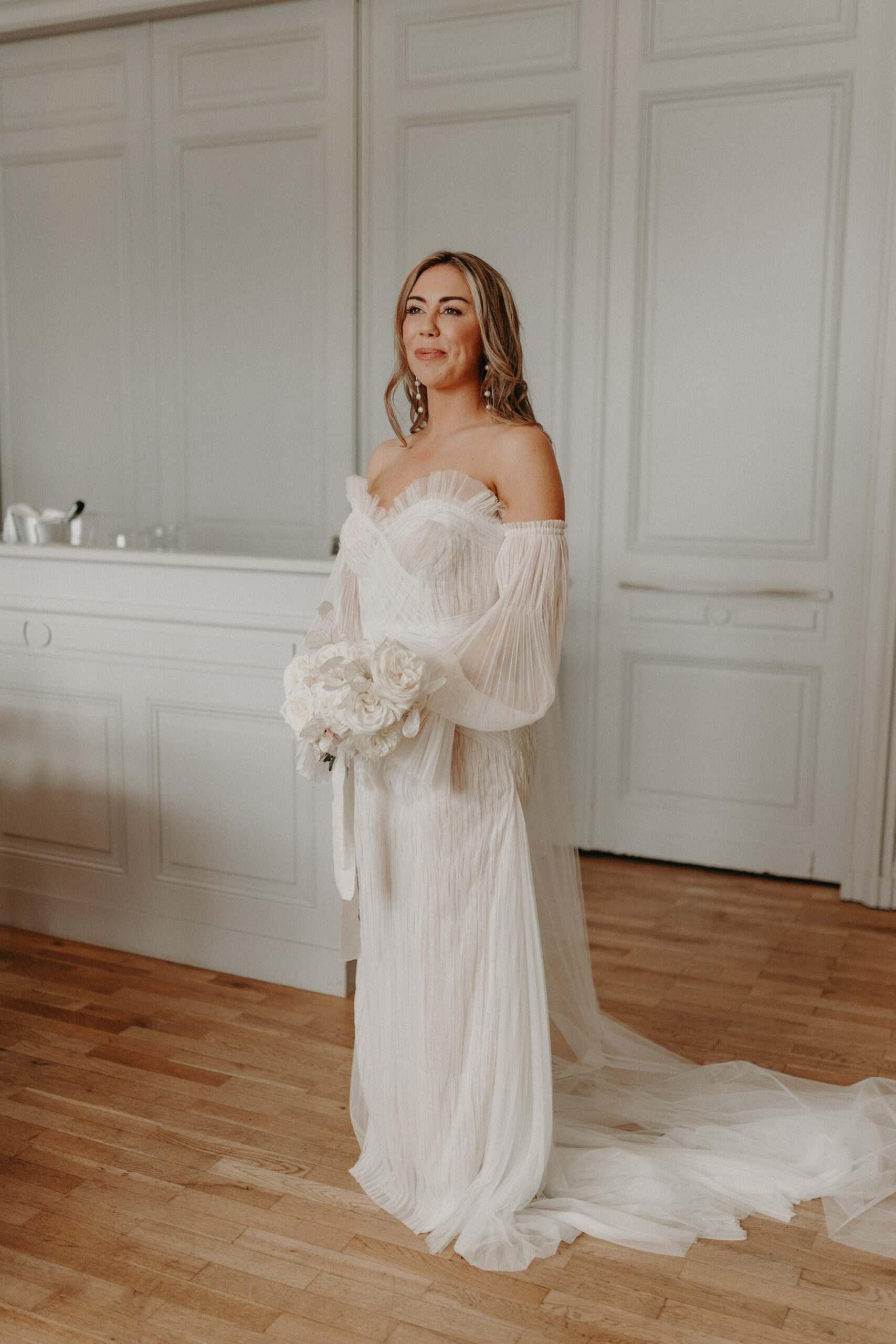 A bride stands alone in a getting-ready portrait inside a room with white wood-panelled walls and warm parquet flooring, likely within a French chateau. She wears an off-the-shoulder white pleated tulle gown with a sweetheart neckline, long sheer draped sleeves, and a floor-length train. She holds a rounded bouquet of white peonies tied with a white ribbon, and wears pearl drop earrings with her hair styled in loose waves. The full-length portrait is shot against the panelled wall, with a vanity and mirror partially visible to the left, suggesting a bridal preparation room.
