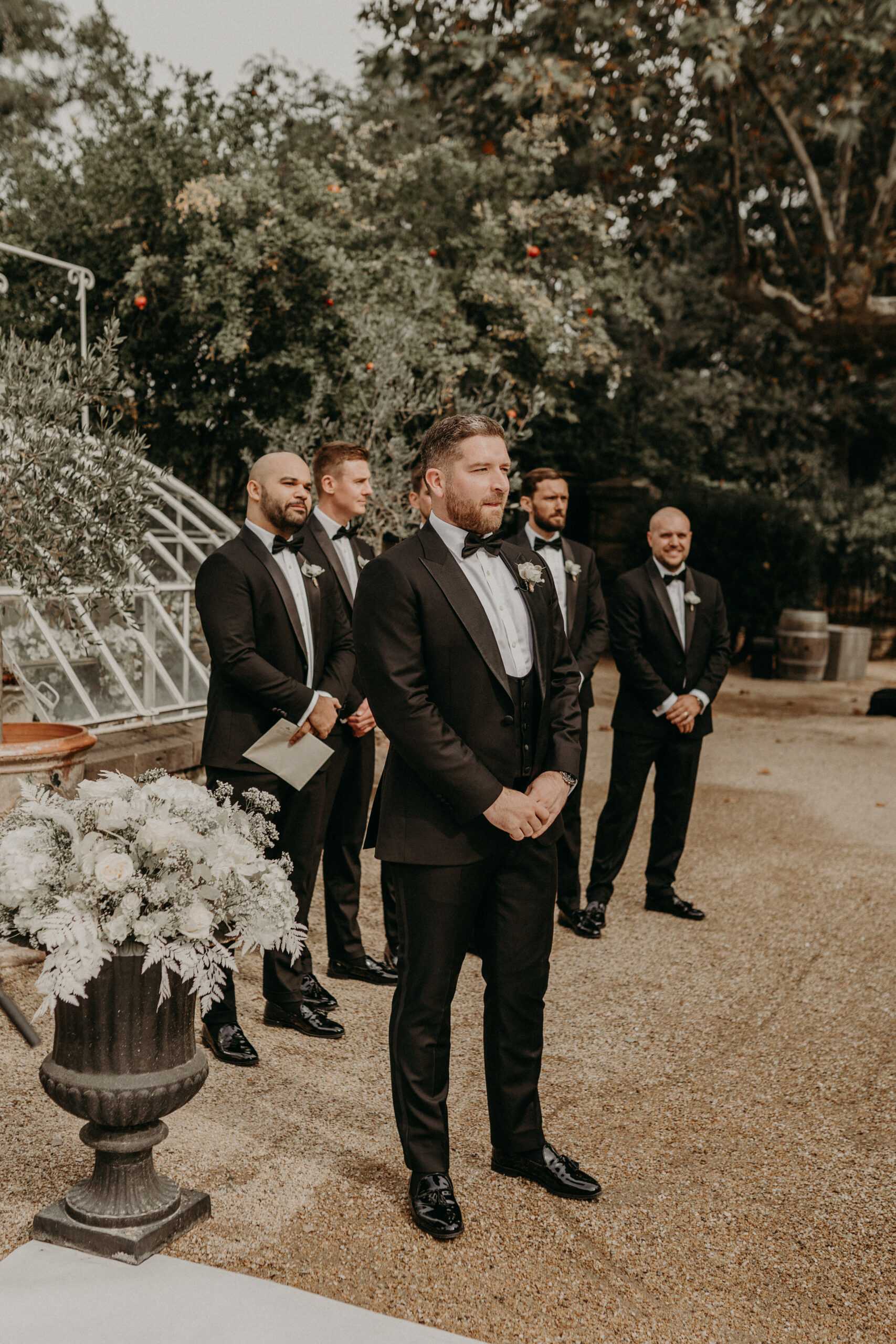An outdoor wedding ceremony moment showing the groom standing at the altar awaiting the bride, with four groomsmen positioned behind him. All five men wear matching black tuxedos with black bow ties, white dress shirts, and black patent leather shoes, each with a white boutonniere. The ceremony takes place on a gravel courtyard, with a large dark cast-iron urn in the foreground filled with white roses, white peonies, and soft silvery-green foliage. The groomsmen stand in pairs behind the groom, one holding a folded program. The setting appears to be a garden or estate grounds with mature trees and what looks like a greenhouse structure visible to the left. The styling is classic and formal. Medium-wide shot taken from a slight low angle.