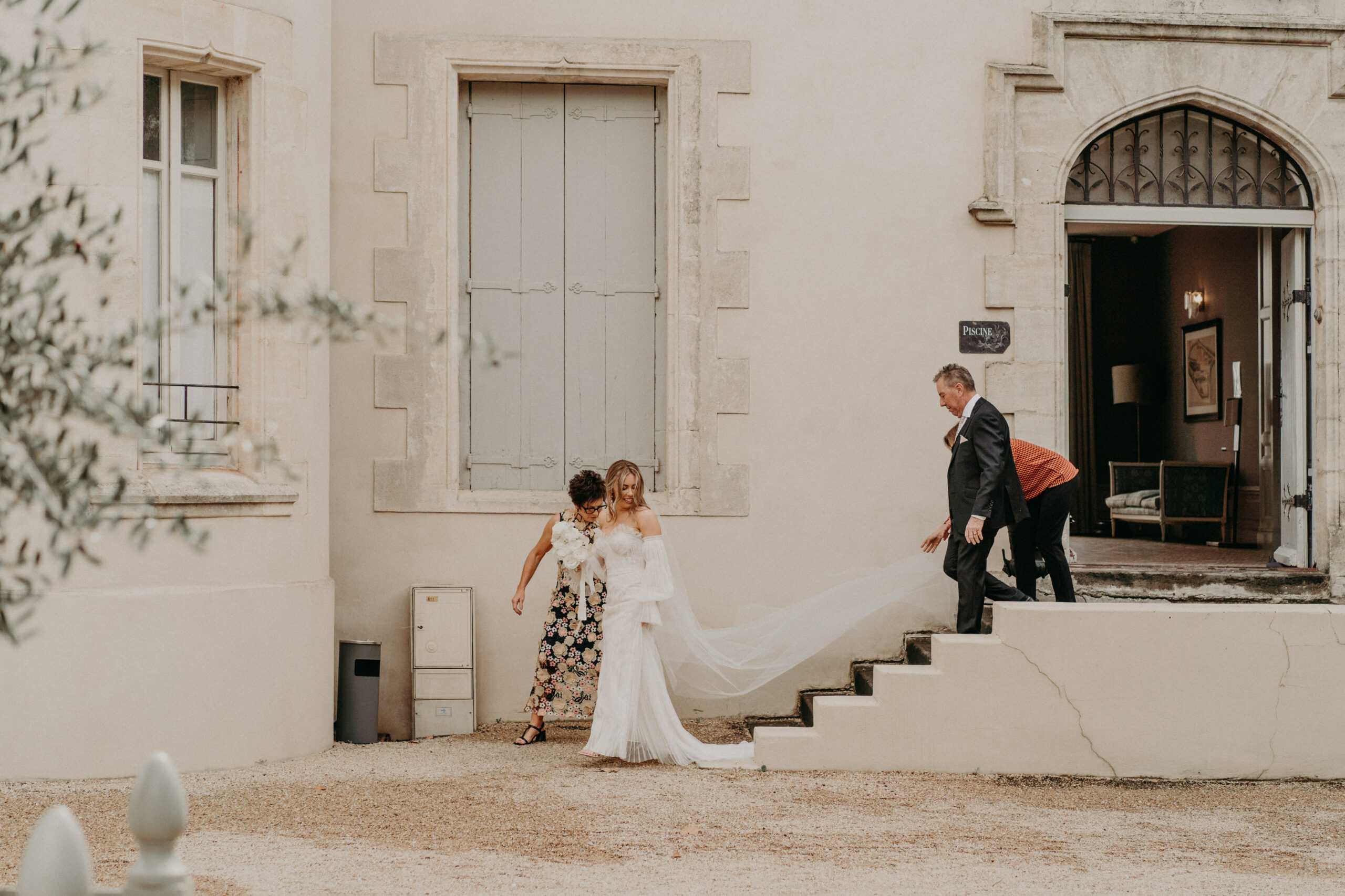 A bride is being helped down exterior stone steps at a French chateau, with two guests assisting her — a woman in a dark floral print dress steadying her from the front and a man in a dark suit behind her. The bride wears an off-the-shoulder white fitted gown with feather or tulle sleeve details and a long flowing veil that trails behind her; she holds a white bouquet with ribbon ties. A third person in an orange top is visible on the steps near the entrance. The chateau facade features pale rendered walls, blue-grey shuttered windows, and a pointed Gothic-arch ironwork doorway with a sign reading 'Piscine,' with an interior room partially visible through the open door. The shot is a wide environmental portrait taken from ground level, framing the figures against the chateau exterior. Potential venue feature image.