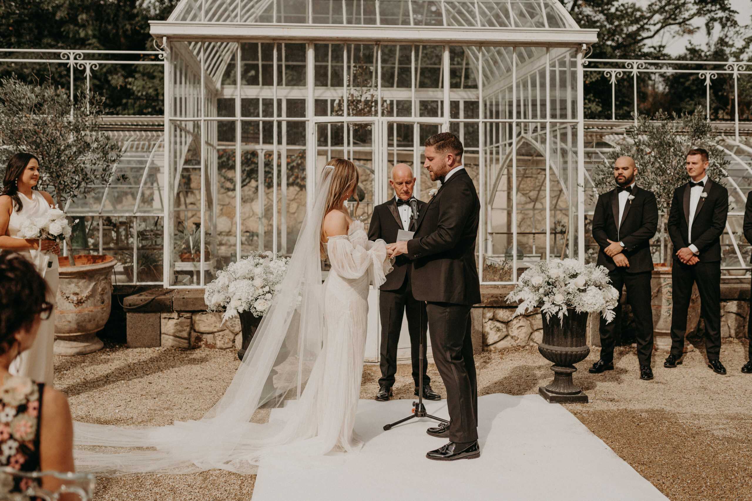 An outdoor wedding ceremony is taking place in front of a white ornate Victorian-style glasshouse greenhouse structure. The bride, wearing an off-shoulder white gown with long billowing sleeves, a cathedral-length veil, and her hair down, faces the groom as they exchange rings or vows; the groom is dressed in a black tuxedo with black bow tie. An officiant in a black suit reads from a booklet behind them at a microphone stand. Two groomsmen in black tuxedos stand to the right, while at least one bridesmaid in a white dress holding a white floral bouquet is visible to the left. Large cast-iron urns filled with voluminous arrangements of white flowers — including what appears to be hydrangeas and gypsophila — flank the altar area on a gravel surface with a white aisle runner. The overall decor palette is black and white with a classic, formal styling. Medium-wide shot taken from behind and slightly to the side of the bride, with guests visible in the foreground.