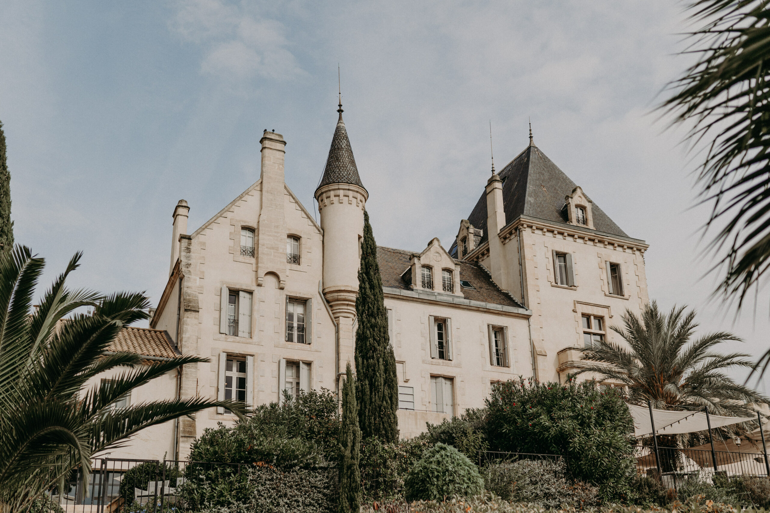 A wide exterior shot looking up at a French chateau featuring cream-colored limestone architecture with dark slate-tiled roofs, a pointed turret, multiple chimneys, and dormer windows across three stories. The building's classical French château style is clearly visible with its symmetrical facades and ornate stonework detailing around the window frames. A white sail shade canopy and what appears to be an outdoor terrace structure are partially visible at the lower right, suggesting an event or hospitality setup. No people are present in the frame. Potential venue feature image.