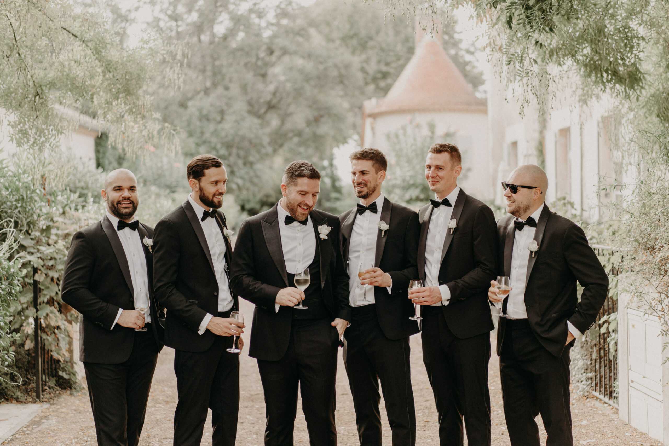 A groomsmen portrait taken outdoors at what appears to be a French chateau estate, with a terracotta-roofed turret and stone buildings visible in the background. Six men are posed casually along a gravel path, all wearing matching black tuxedos with black bow ties, white dress shirts, and white boutonnières, with one man wearing black sunglasses. Each man holds a champagne or wine glass, and the group is laughing and conversing in a relaxed, candid manner. The shot is a medium-wide portrait with the group arranged in a loose line facing slightly inward toward each other.
