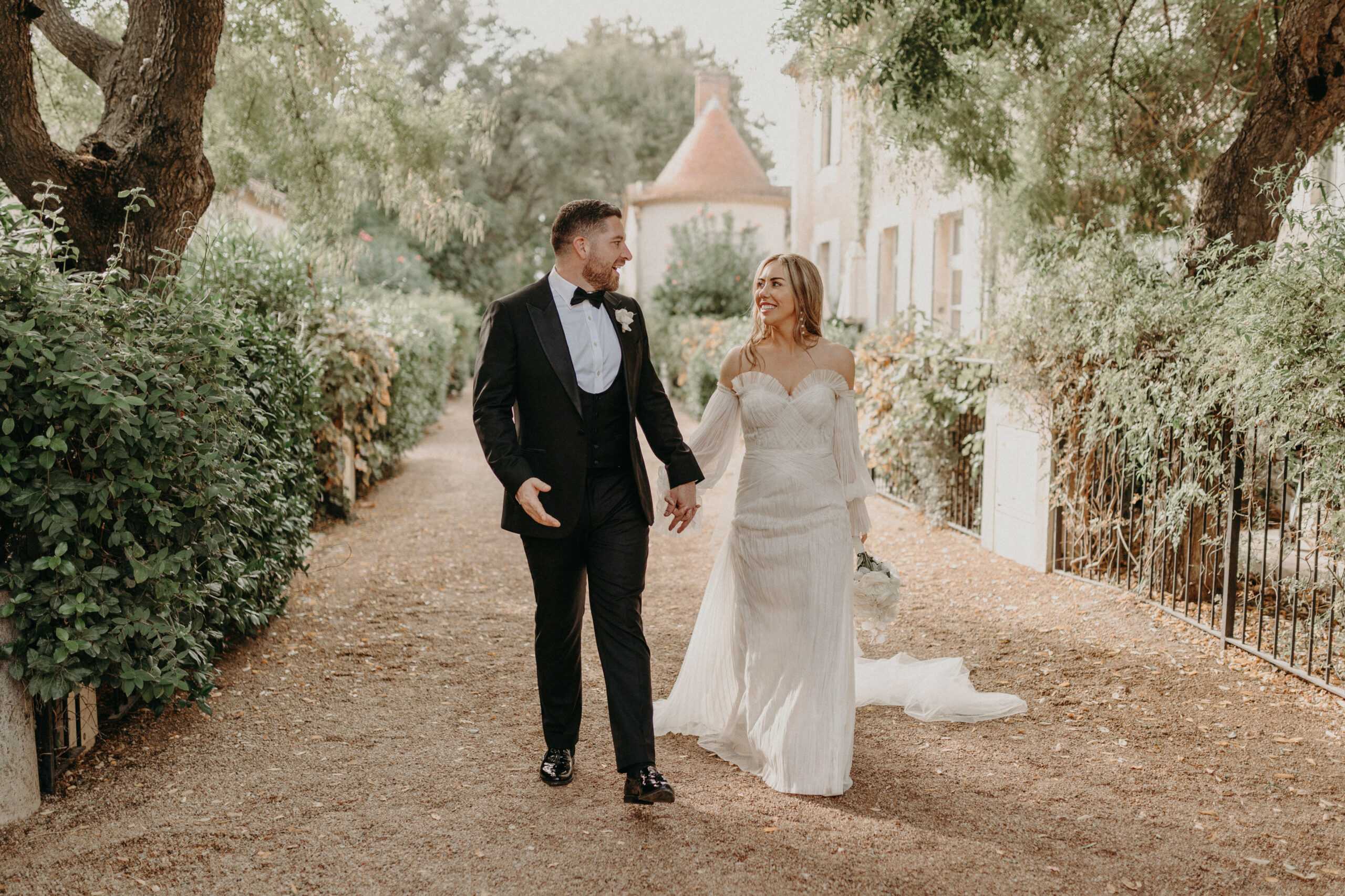 A couple portrait taken outdoors on a gravel path lined with hedges and mature trees, with a stone chateau building visible in the background. The bride wears a fitted ivory off-the-shoulder gown with delicate beading or pleating detail and flowing off-the-shoulder long sleeves, carrying a small white bouquet at her side, while the groom wears a black tuxedo with bow tie, white dress shirt, and a white boutonniere. The two are walking hand-in-hand toward the camera, smiling at each other. The image is a medium full-length shot with warm, soft natural light and a classic-meets-romantic styling aesthetic.