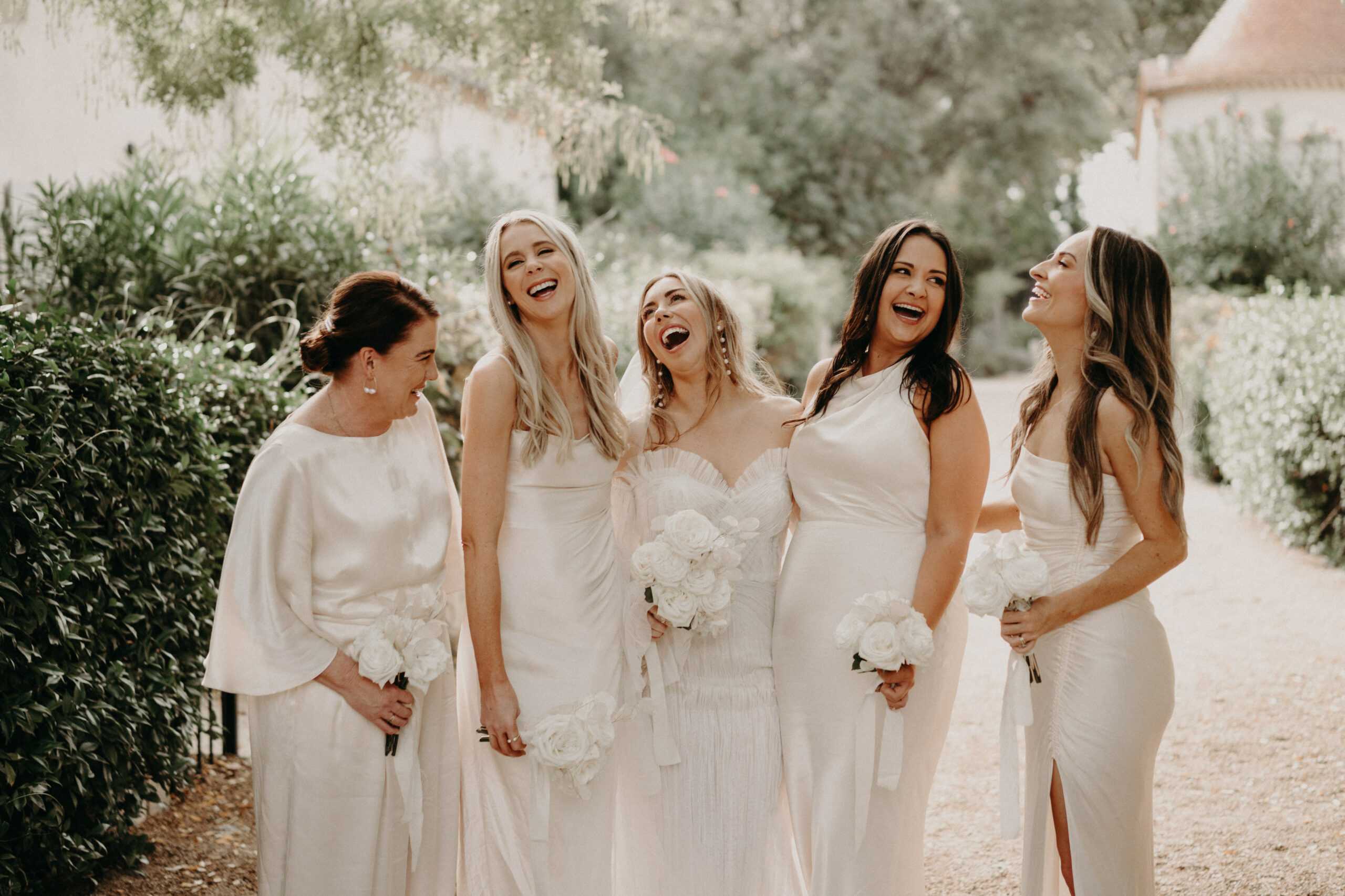 A bridal party portrait taken outdoors on a gravel path, likely within a French estate or chateau garden. The bride stands in the center wearing a strapless sweetheart neckline gown with a pleated bodice and sheer long sleeves, holding a large bouquet of white roses with a white ribbon tie. She is flanked by four bridesmaids, all dressed in varying styles of ivory and white floor-length gowns — including a cape-sleeve satin style, a slip-style halter dress, a strapless column gown, and a satin strapless dress with a side slit — each carrying a smaller bouquet of white roses. The overall color palette is entirely white and ivory, creating a monochromatic bridal party look. All five women are laughing openly, caught mid-movement in a candid moment. The shot is a medium wide portrait with soft, warm natural light.