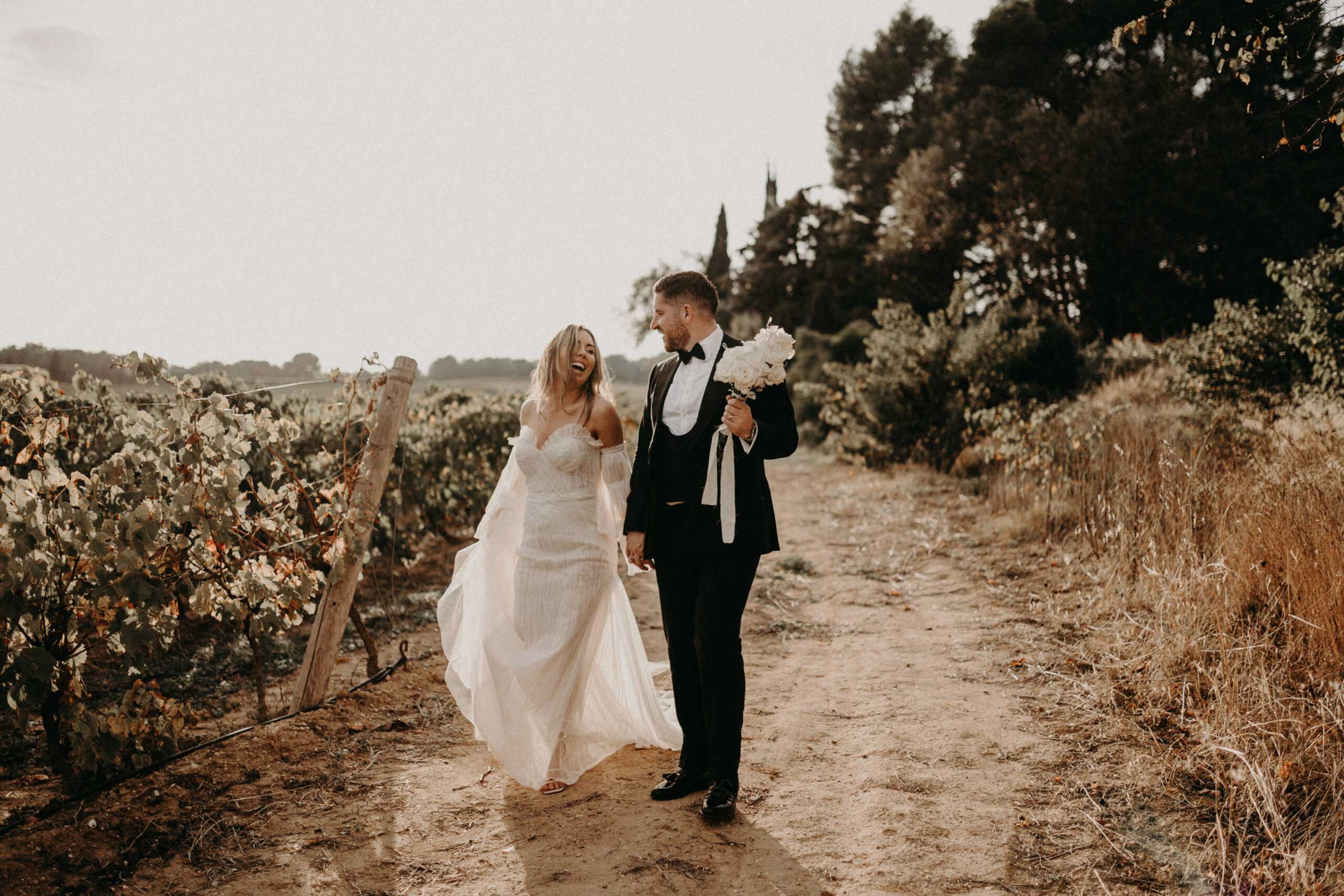 A couple portrait taken outdoors along a dirt path running beside a vineyard. The bride wears an off-the-shoulder ivory gown with a flowing, textured skirt and detached sleeves, and is laughing while holding the groom's hand. The groom wears a black tuxedo with a white dress shirt, black bow tie, and black dress shoes, and carries a bouquet of white peonies with white ribbon trailing from his hand. Both are walking toward the camera in a relaxed, candid moment. The overall styling is classic with a relaxed, natural edge. Wide shot with warm, muted tones consistent with late afternoon natural light.