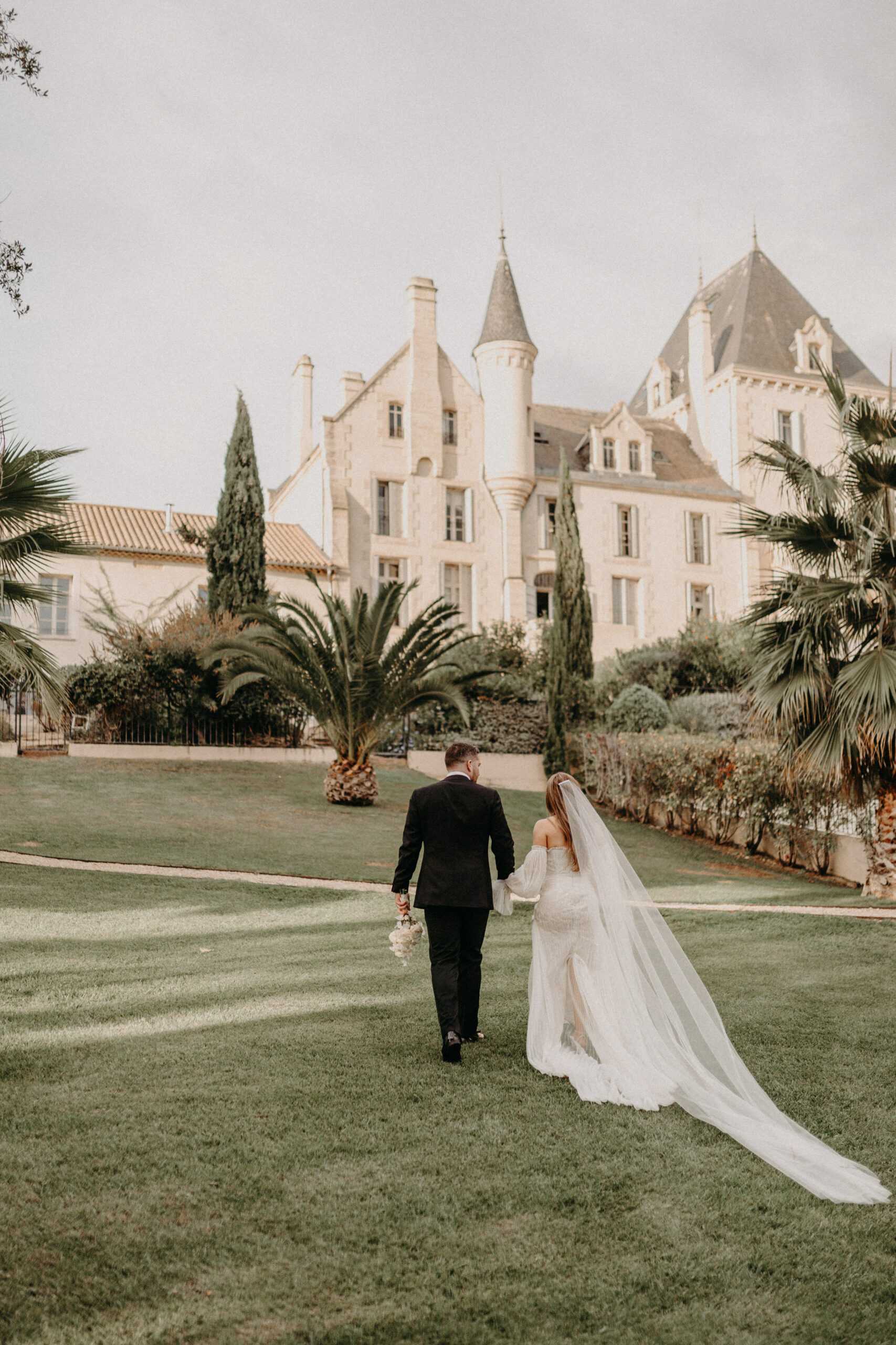 A couple walks hand-in-hand across a manicured lawn toward a large French chateau with limestone architecture, pointed turrets, and slate-grey roofing. The bride wears an off-the-shoulder white gown with long sleeves and a dramatic cathedral-length veil that trails behind her across the grass; she carries a small bouquet with cream and blush tones. The groom wears a dark navy or black suit. The shot is taken from behind at a medium-wide distance, with the chateau centered in the background framed by cypress trees and palm trees. Potential venue feature image.