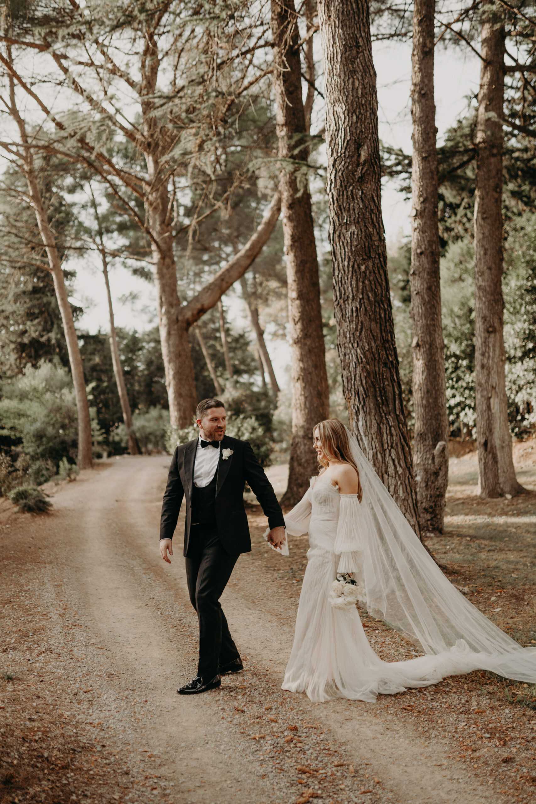A couple portrait shot outdoors on a gravel path lined with tall pine trees. The groom, dressed in a black tuxedo with bow tie and a white boutonniere, walks slightly ahead while holding the bride's hand and looking back at her. The bride wears an off-the-shoulder white gown with a flowing tulle cape or dramatic veil that trails behind her, and she carries a small bouquet of white peonies. The styling is modern classic with a relaxed, editorial feel. The image is taken as a full-length portrait with natural afternoon light filtering through the trees.
