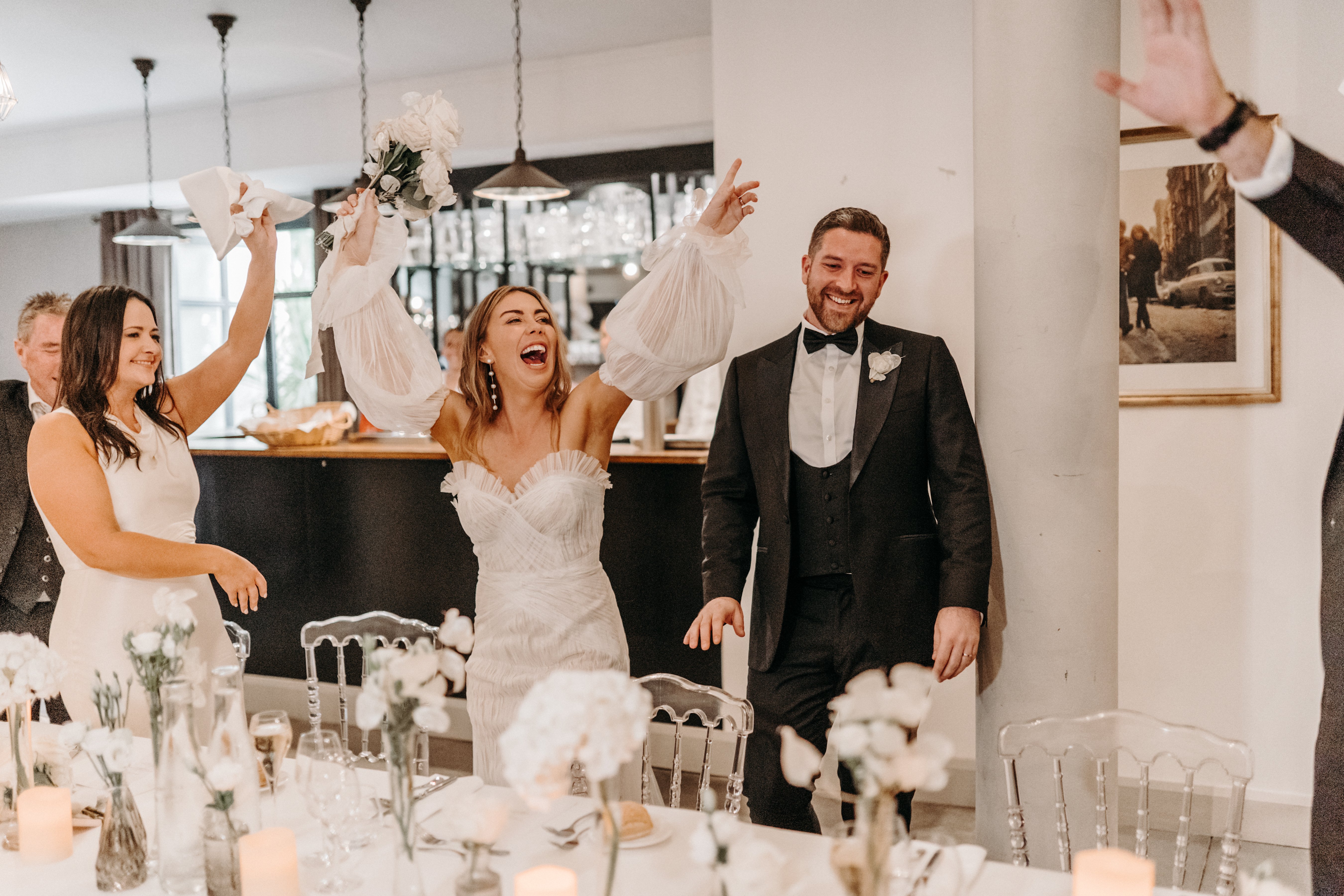 The bride and groom are making their entrance into an indoor reception room, greeted with cheers from guests. The bride wears a strapless ruched white gown with a separate sheer puff-sleeve bolero and holds her white floral bouquet raised in the air while laughing openly; the groom wears a black tuxedo with a three-piece suit, bow tie, and a white floral buttonhole. A female guest in a white sleeveless dress stands beside them clapping, and at least two other guests are partially visible. The reception table in the foreground is styled with a white and green palette featuring small bud vases of white roses and greenery, taper candles, pillar candles, and clear acrylic ghost chairs. The room has an interior bar area with pendant lighting and framed black-and-white photography prints on the walls, giving a modern, clean aesthetic. Medium shot taken from a low angle beside the reception table.