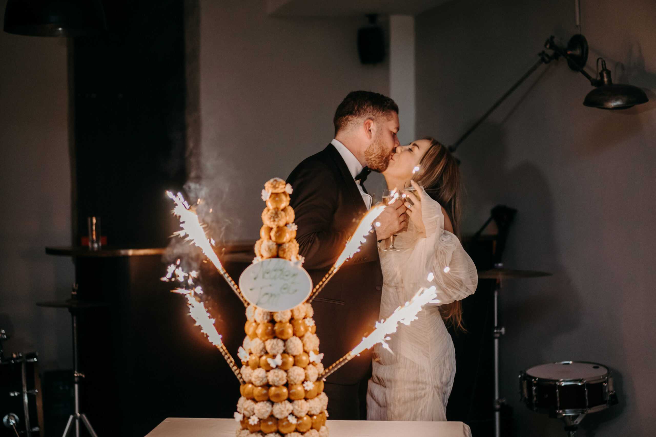 The couple shares a kiss during the croquembouche presentation at an indoor reception, with the bride holding a champagne flute. The pièce montée — a tall tower of caramel-glazed choux puffs decorated with small white sugar flowers and a personalized oval plaque — is fitted with lit sparklers that radiate dramatically outward. The groom wears a black tuxedo with a bow tie, and the bride wears a white long-sleeve dress with a sheer, textured overlay. The venue has an industrial-modern aesthetic with dark grey walls, exposed structural columns, wall-mounted metal arm lamps, and a drum kit visible in the background, suggesting a live band was part of the evening. The shot is a medium portrait framing the couple and dessert together, with warm sparkler light contrasting against the dim, moody room.