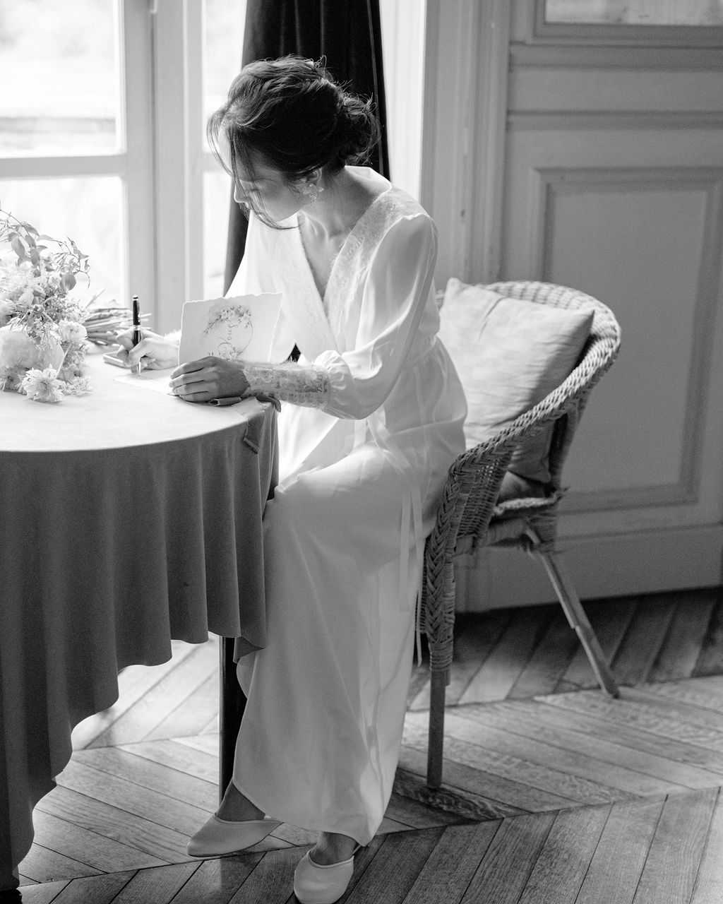 This black-and-white image shows a bride seated at a small round draped table beside a large window, writing in what appears to be a wedding card or vow booklet with illustrated floral detailing on the cover. She is wearing a long-sleeved, floor-length bridal gown with lace cuff detail and flat bridal slippers, with her dark hair styled in a loose updo. A bouquet of flowers rests on the table beside her, and she is seated in a wicker chair with a cushion; classic wood paneling and herringbone parquet flooring are visible in the background, suggesting an interior room of a French chateau or manor house. The image is a full-length portrait shot with soft, high-contrast natural window light, giving it bright highlights and deep mid-tones.